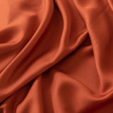 The Linenly Bamboo Fitted Sheet in Copper Sunset is captured close-up, displaying its smooth texture and gentle folds. Its rich hue and luxurious sheen exude sophistication and comfort, reminiscent of rust-colored silk fabric.