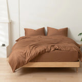 A neatly made bed features Linenly's Bamboo Quilt Cover Set in Terracotta and two pillows on a light wooden frame in a minimalistic room with pale walls and flooring. A small plant is visible on the right side of the image.