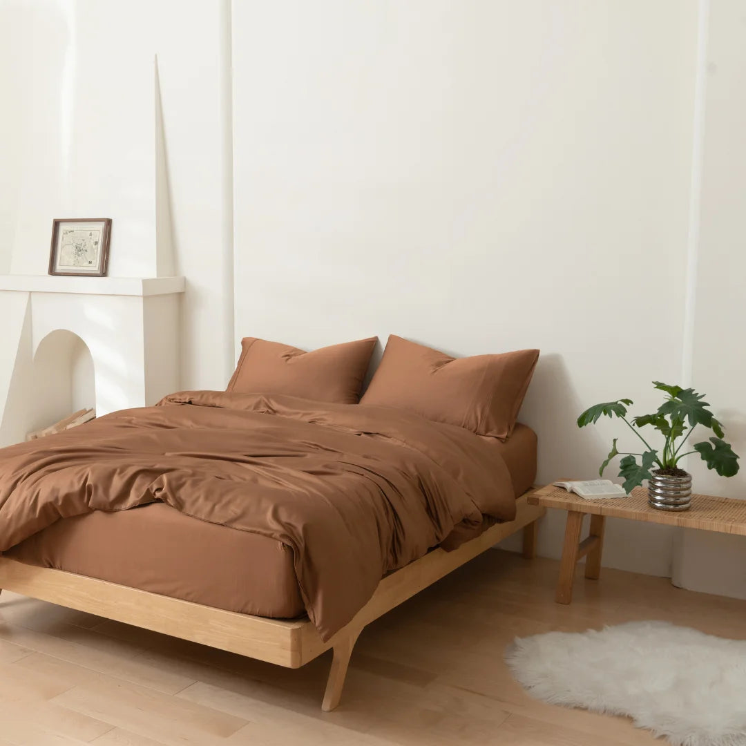 A minimalist bedroom with a wooden bed frame and brown bedding, featuring the Linenly Bamboo Quilt Cover Set in Terracotta, two pillows, a small plant on a wooden bench, a white rug, and a simple white fireplace with a framed picture.