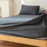 A hand pulls the Linenly Luxe Cotton Sateen Fitted Sheet in Charcoal over a wooden bed corner with matching pillowcases, in a bright, minimalist bedroom with modern bedding.