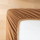 A close-up of the Linenly Luxe Cotton Sateen Fitted Sheet in terracotta being pulled over a white mattress on a light wooden floor, adding a cozy touch to the bedding.