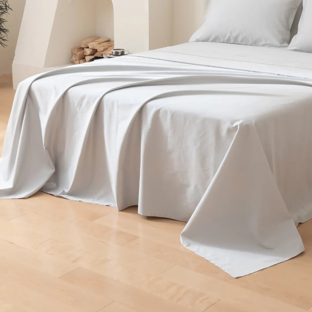 A neatly made bed features the Luxe Cotton Sateen Flat Sheet in Silver by Linenly and white pillows, with part of the sheet elegantly draped on a light wooden floor in a minimal, cozy room. Firewood is stacked on a shelf in the background.