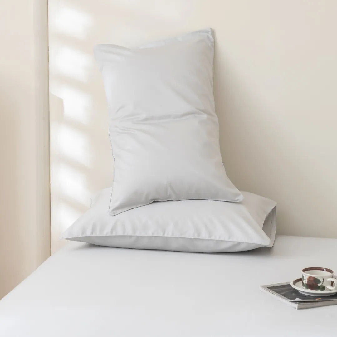 Two white pillows rest on a neatly made bed with the Linenly Luxe Cotton Sateen Flat Sheet in Silver. A cup and saucer sit on a tray nearby as soft natural light casts gentle shadows across the room.