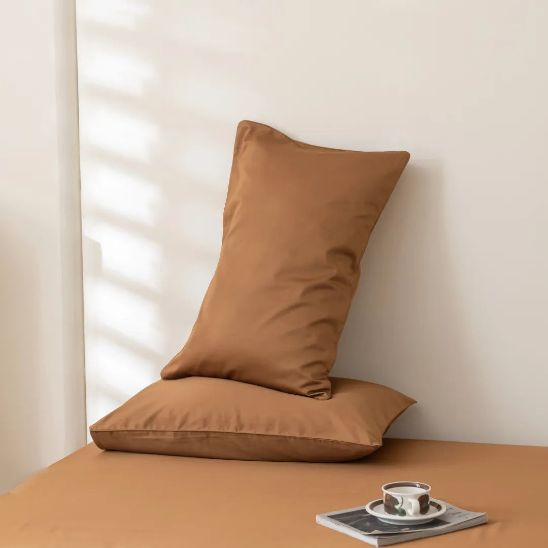 Two brown pillows rest against a light wall; one leans on the other, which lies flat on the Linenly Luxe Cotton Sateen Flat Sheet in Terracotta. A cup, saucer, and magazine are nearby as soft light casts gentle shadows on the wall.