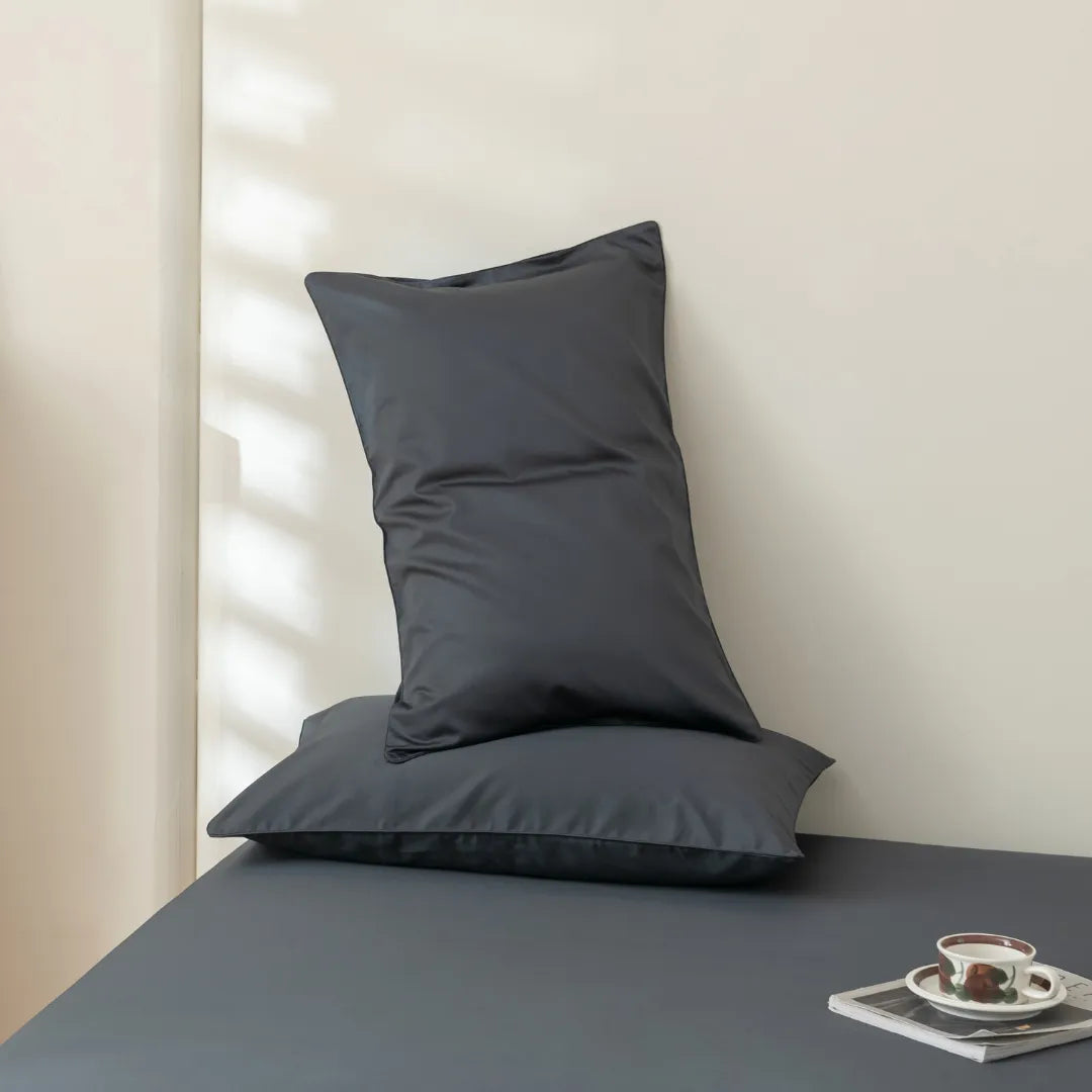 Two Linenly Luxe Cotton Sateen Pillowcases in charcoal are stacked on a matching gray bed. Sunlight casts striped shadows on the cream wall, while a cup and saucer sit on a small tray in the bed’s corner.
