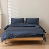 A neatly made bed features Linenly's Luxe Cotton Sateen Quilt Cover in Midnight and two matching pillows, resting on a light wooden frame in a minimalistic room with pale walls and wooden flooring.