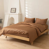 A bright, minimalist bedroom features a neatly made bed with Linenly’s Luxe Cotton Sateen Quilt Cover in Terracotta and two pillows on a light wood frame, set against white walls with a fireplace and framed picture on the mantel.