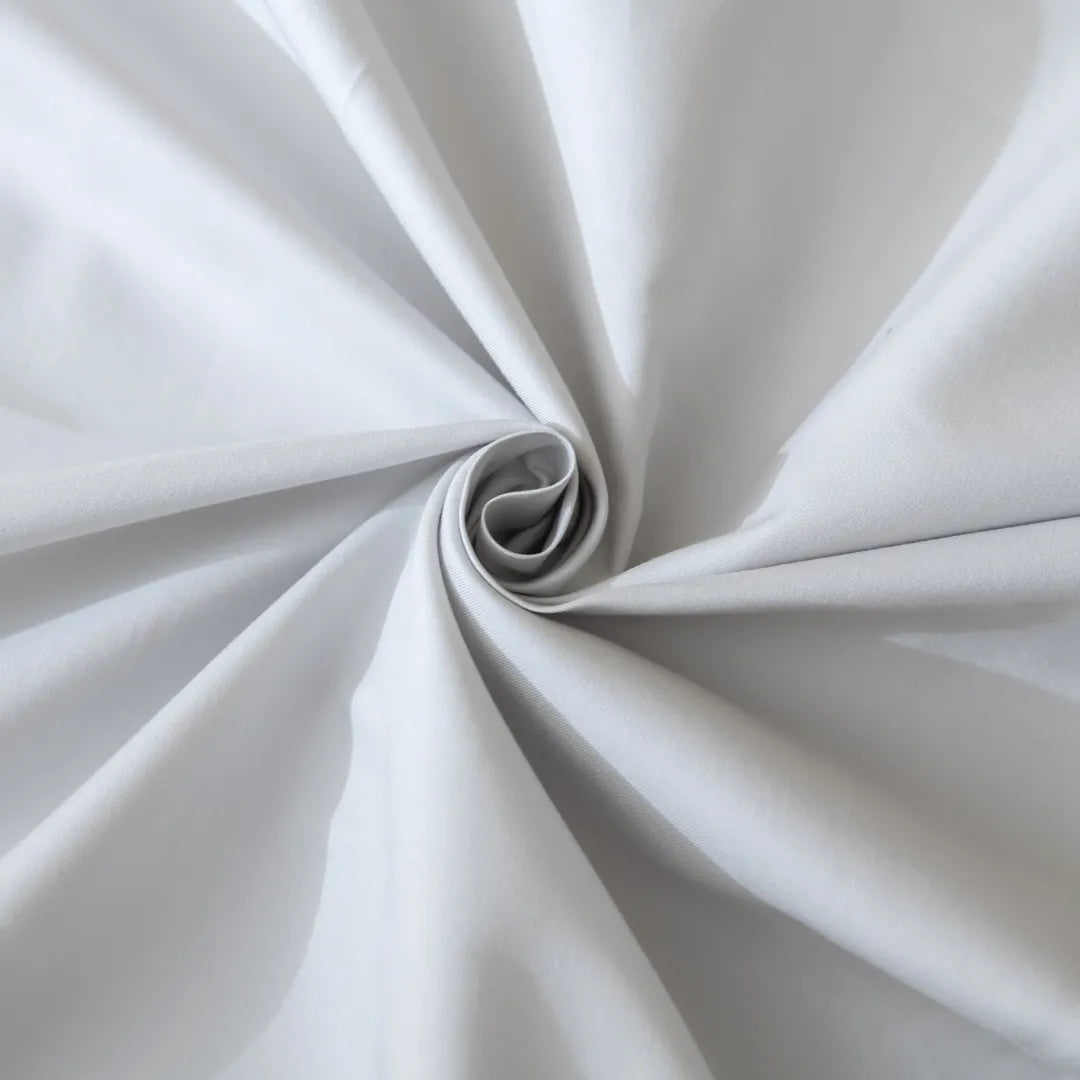 A close-up of Linenly’s Luxe Cotton Sateen Sheet Set in Silver showcases the light gray fabric, gathered and twisted into a spiral to reveal silky folds and soft shadows in its smooth sateen weave.