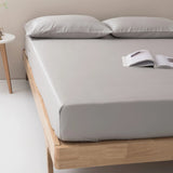 A neatly made bed features the Linenly Bamboo Fitted Sheet Set in Silver and two pillows, with an open book on top. The wooden-framed bed sits beside a small white round side table, enhancing the room's sustainable luxury.