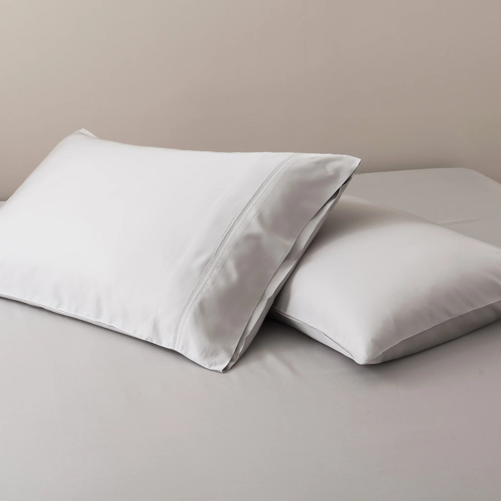 Two white pillows with Linenly’s Bamboo Pillowcase Set in Silver rest on a neatly made bed with light gray sheets against a plain beige wall.