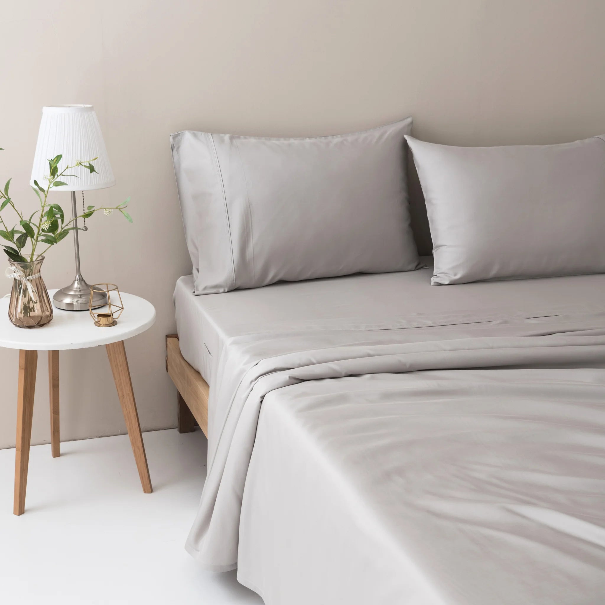 A minimalist, modern bedroom features a neatly made bed with Linenly’s Bamboo Pillowcase Set in Silver, next to a small round white nightstand topped with a lamp, glass, book, and a vase of green branches.