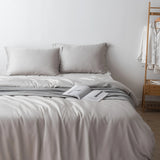 A neatly made bed features the Linenly Bamboo Quilt Cover Set in Silver with two pillows, a striped blanket at the foot, and an open book. In the background, a wooden rack displays white pajamas against a plain white wall.