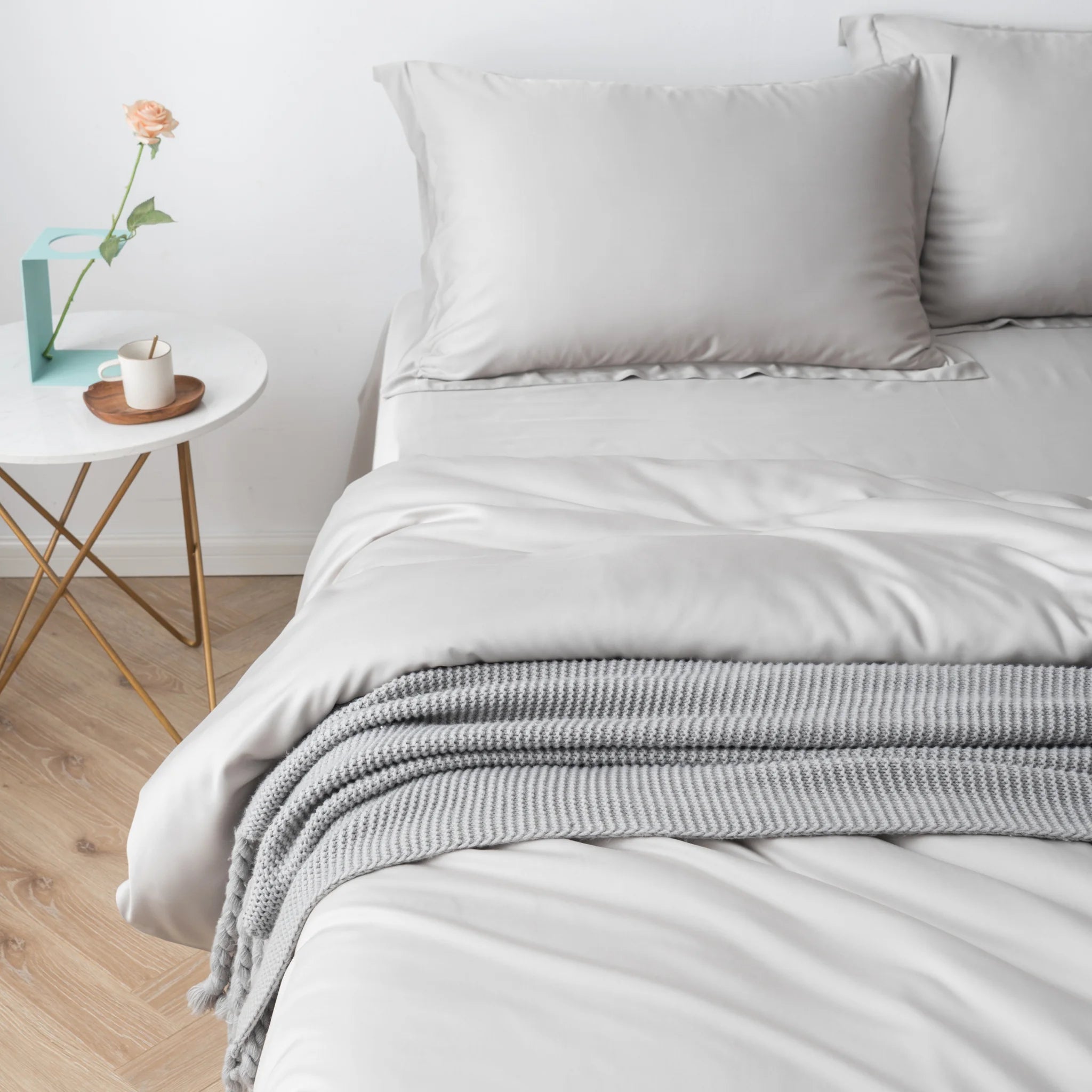 A neatly made bed features Linenly’s Bamboo Quilt Cover Set in Silver, topped with two pillows and a folded gray knit blanket on the cooling cover. Beside it, a round white table holds a cup, saucer, book, and a peach rose in a vase.