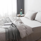 A bed made with Linenly's Bamboo Quilt Cover Set in Silver and a knit blanket, topped with an open book. Next to it, a round table holds a potted flower, mug, and saucer. Soft daylight streams through sheer curtains.