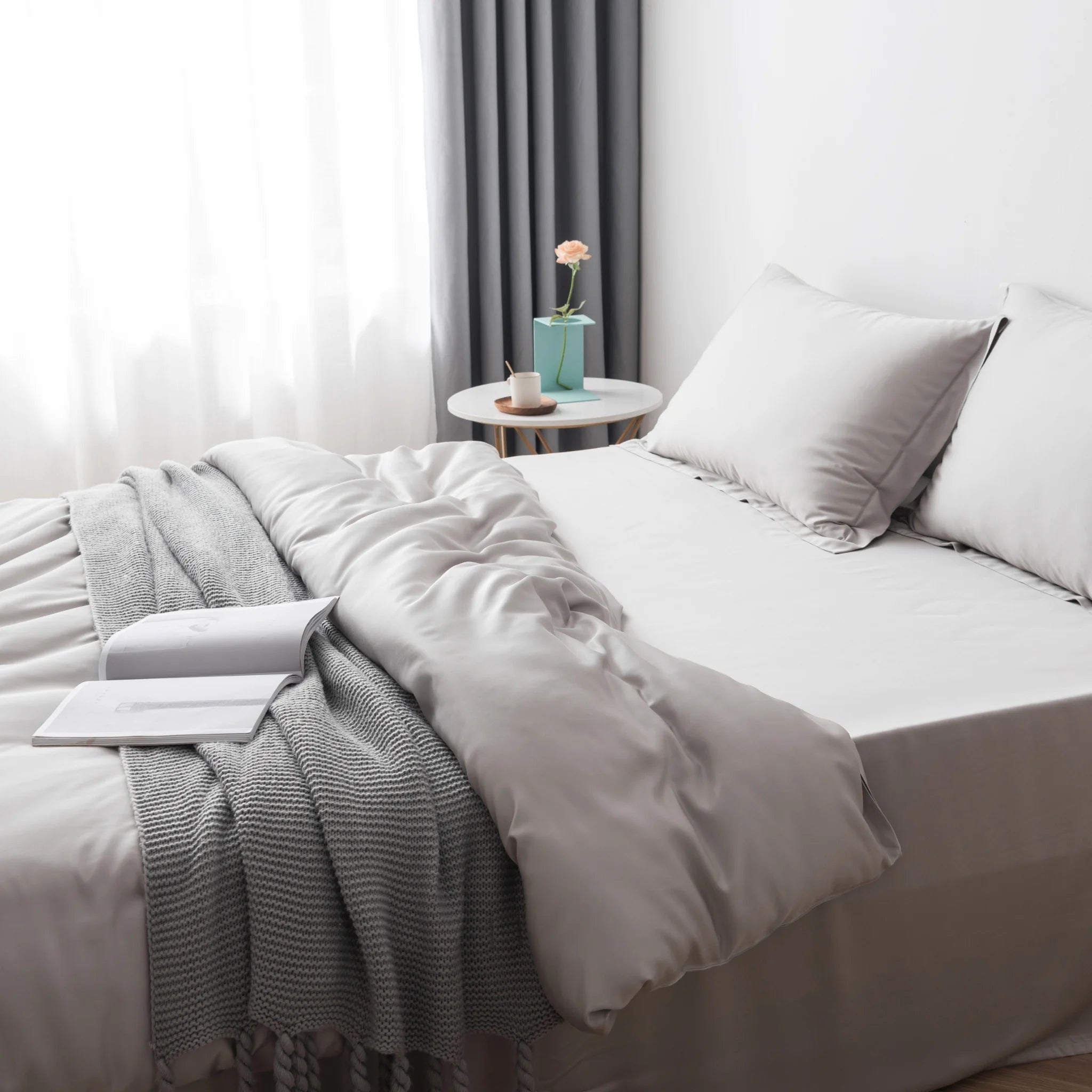 A bed made with Linenly's Bamboo Quilt Cover Set in Silver and a knit blanket, topped with an open book. Next to it, a round table holds a potted flower, mug, and saucer. Soft daylight streams through sheer curtains.